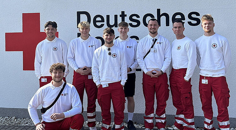 Gruppe von acht Personen in weiß-roter Dienstkleidung mit Rotkreuz-Emblem vor einer Wand mit der Aufschrift 'Deutsches R...'.