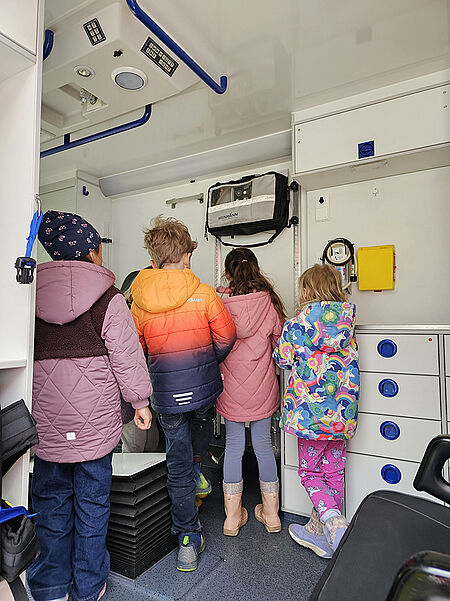 Vier Kinder in bunter Winterkleidung stehen in einem Fahrzeuginnenraum eines Rettungswagens mit weißen Schränken und blauen Griffen